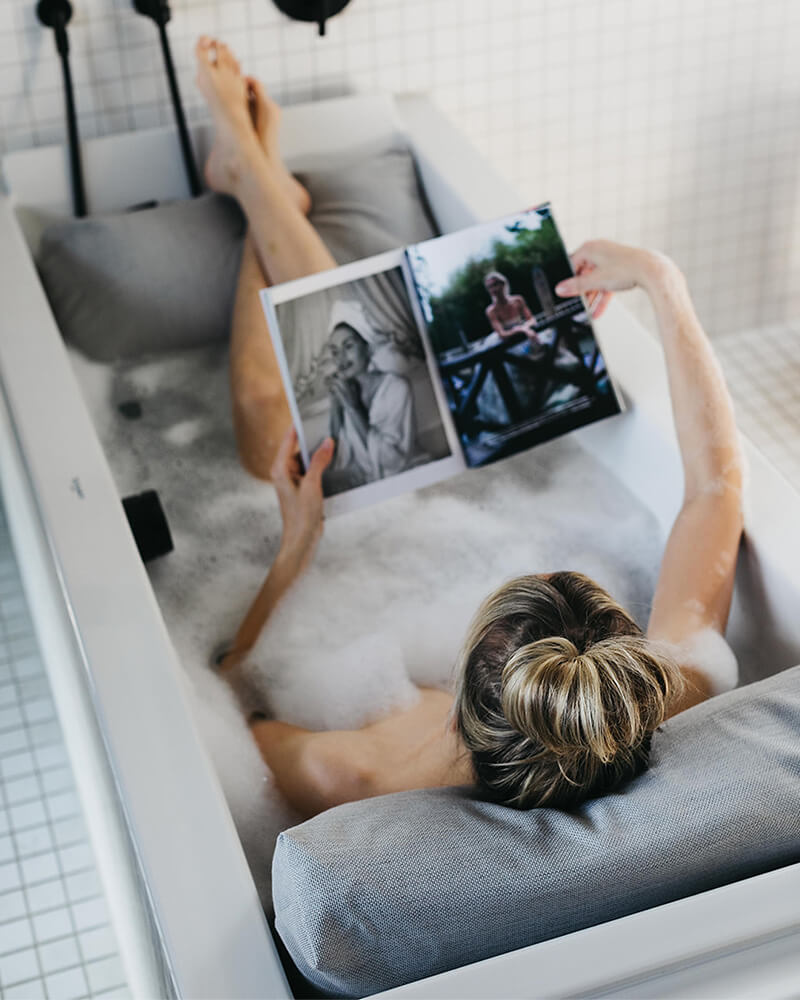 Frau badet mit Rückenkissen (Medium) und Fusskissen in luxuriöser freistehender Badewanne. Rücken, Kopf und Füsse ruhen entspannt auf Badesofas (Farbe Grau). $ Innenform eckig | Size Medium Set | Grau