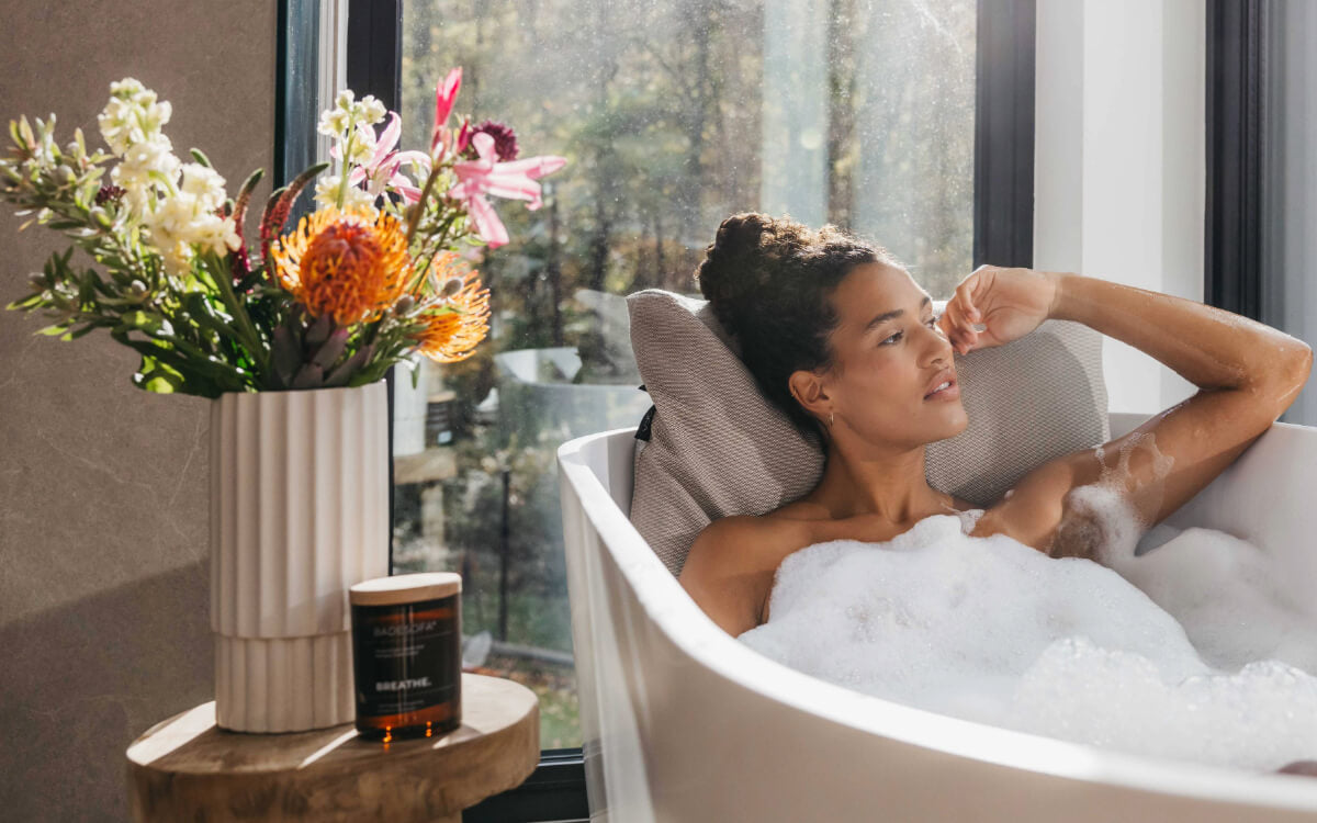 Eine Frau liegt entspannt in einer freistehenden Badewanne, sie lehnt sich an ein Badesofa Rückenkissen Large in der Farbe Beige.