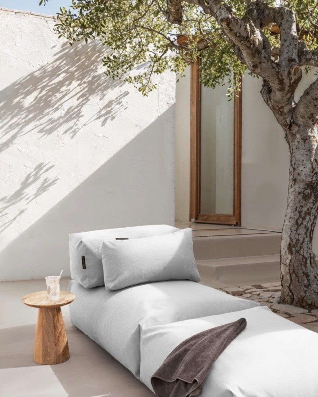 Unter einem Baum steht ein Poolsofa Singel in Grau, daneben ein kleiner Holztisch. Das Rückenkissen und Relaxkissen sind auf dem Poolsofa drapiert.