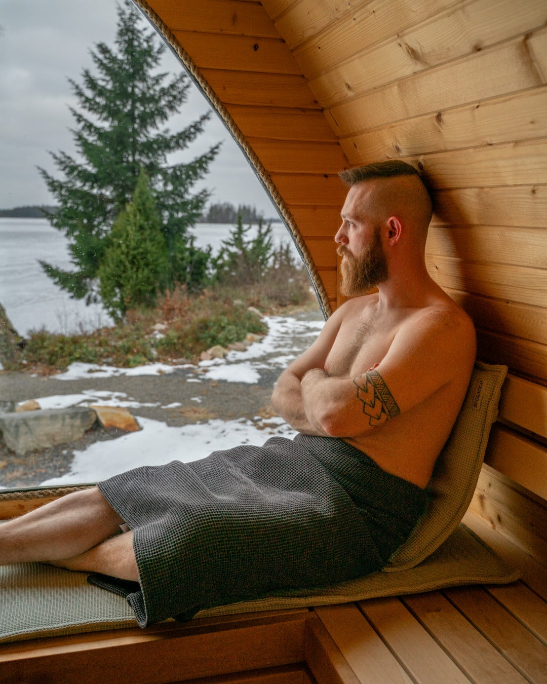 Mann sitzt angelehnt in ein Saunatuch aus Wafflepiqué gewickelt in der Sauna und blickt entspannt aus em Fenster in die Natur.