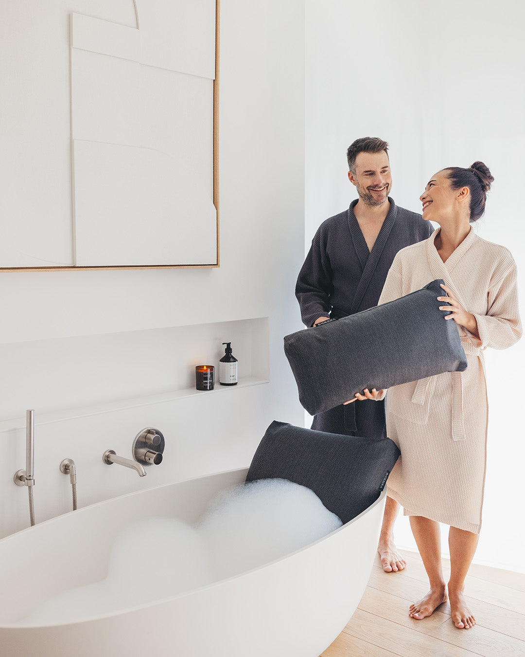 Ein Paar steht vor der Badewanne, beide tragen Bademäntel (er: Anthrazit, sie: Perlmutt), beide lächeln sich an. Die Frau hält ein Badesofa Fusskissen in der Hand, in der Badewanne ist bereits Wasser und ein Badesofa Rückenkissen in Größe Large in Anthrazit.