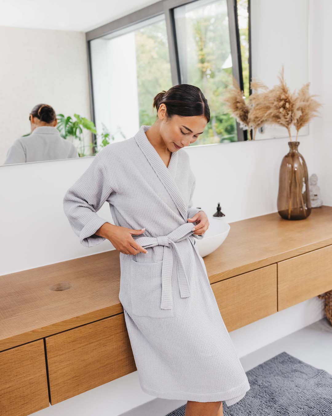 Frau steht im Bademantel (Farbe Hellgrau) angelehnt ans Waschbecken und bindet die Schleife ihres Bademantels.