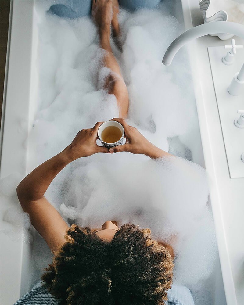 Frau badet mit Rückenkissen (Large) und Fusskissen in luxuriöser freistehender Badewanne. Rücken, Kopf und Füsse ruhen entspannt auf Badesofas (Farbe Blau). $ Innenform eckig | Size Medium Set | Blau